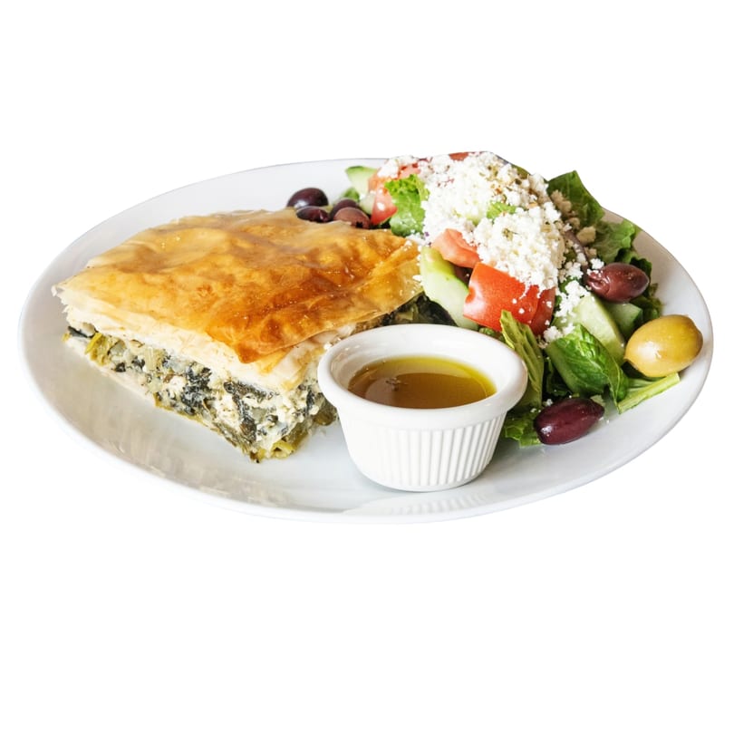 A slice of spanakopita with a side of Greek salad and dressing on a white plate.