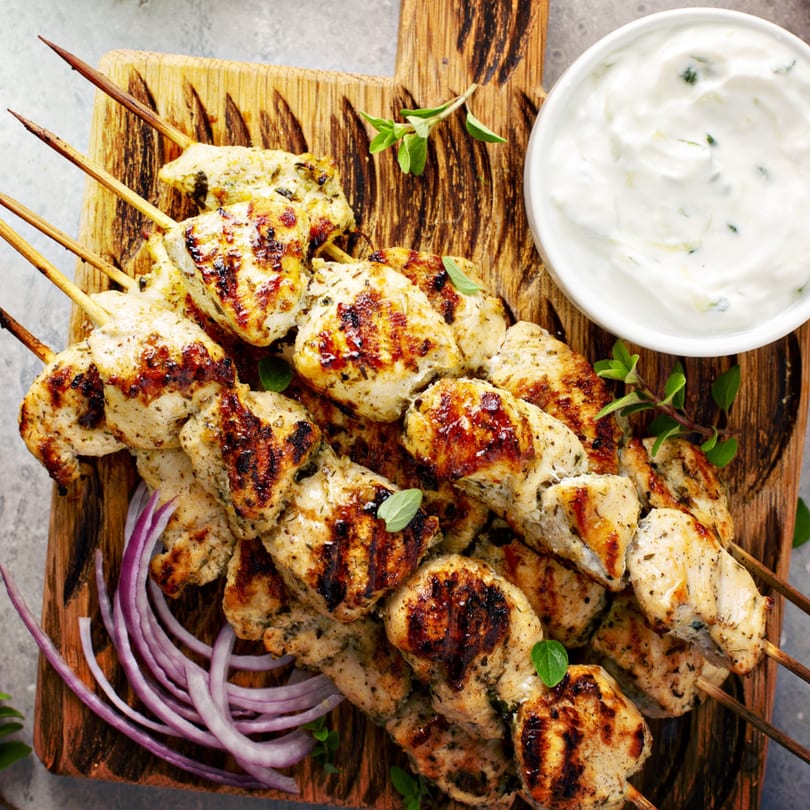 Grilled chicken skewers with tzatziki sauce, red onions, and fresh oregano on a wooden board.