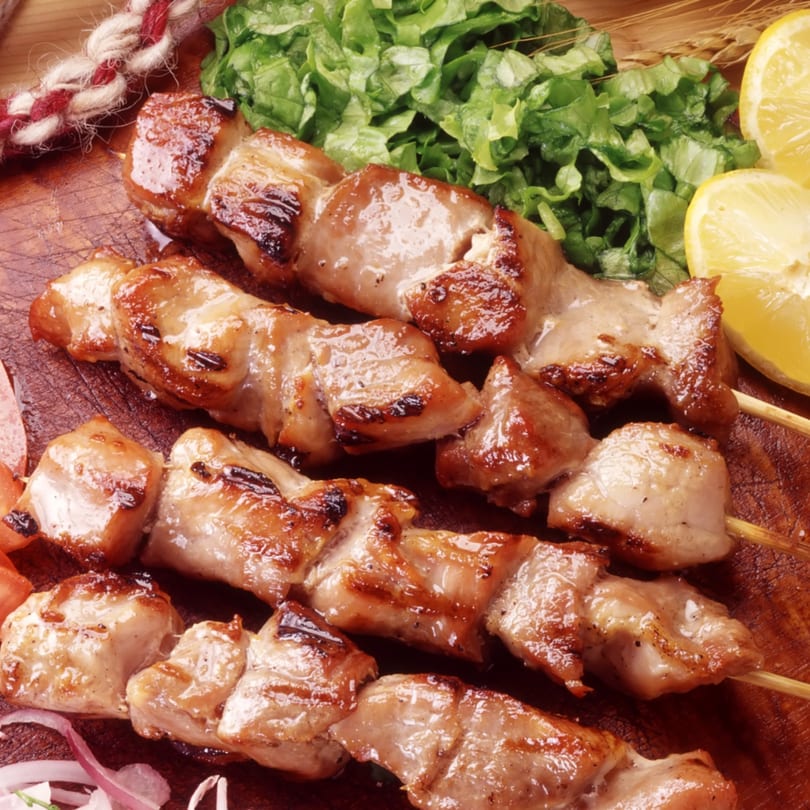 Three grilled chicken skewers with lettuce, lemon, and red onion on a wooden board.