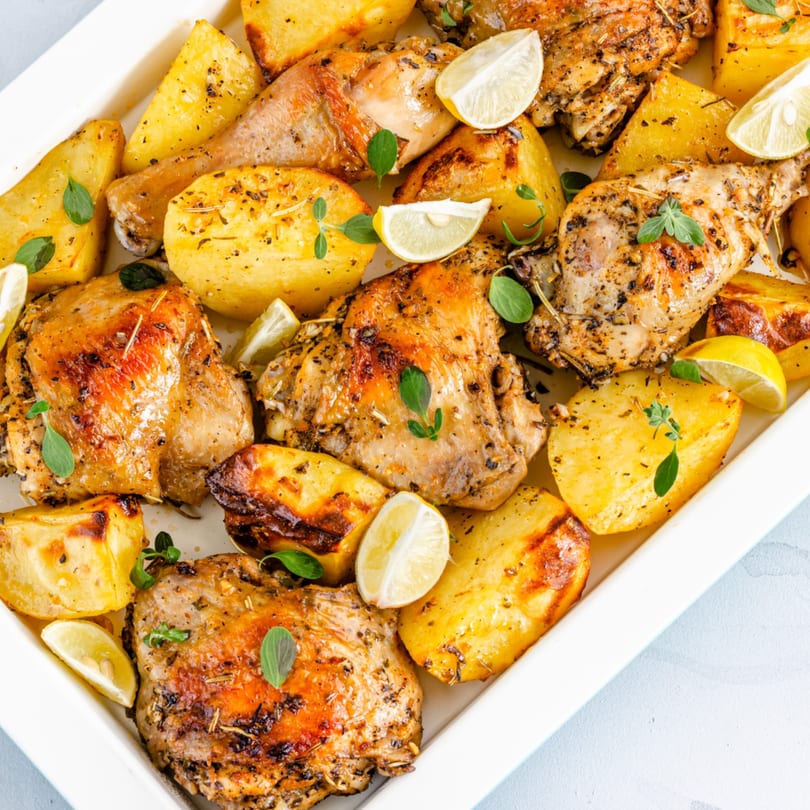 Roasted chicken pieces and potato wedges with lemon slices and herbs in a white baking dish.