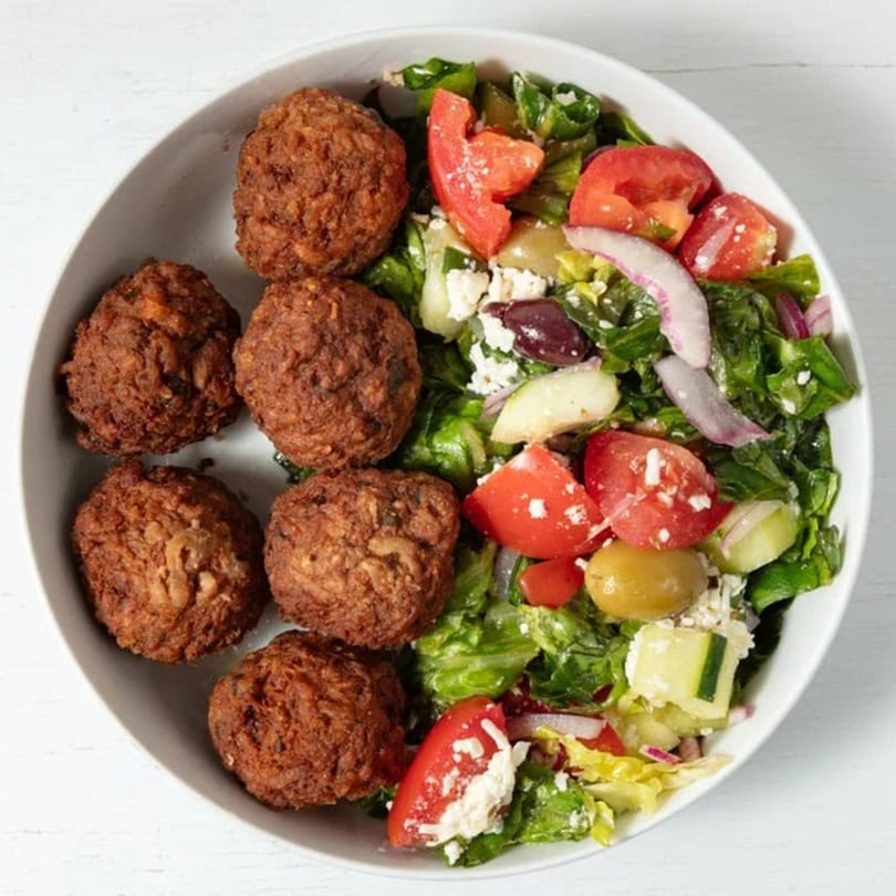 A white bowl filled with golden-brown falafel and a fresh Mediterranean salad with feta, olives, and tomatoes.