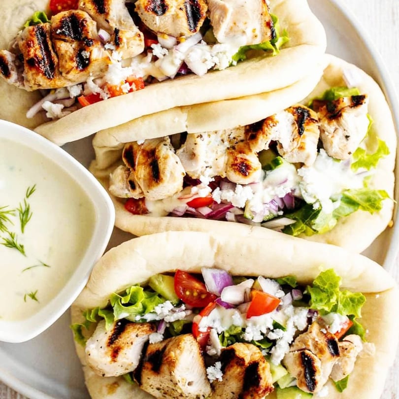 Three Mediterranean chicken gyros with grilled chicken, feta, lettuce, tomatoes, and red onions, with a side of tzatziki.